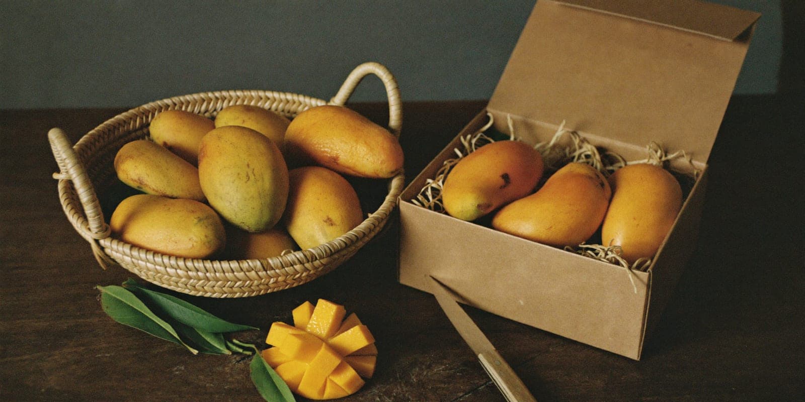An open kraft box beside a basket of ripe mangoes and a freshly cut fruit